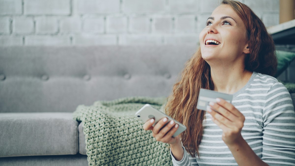 Happy woman holding a credit card and smartphone on the sofa