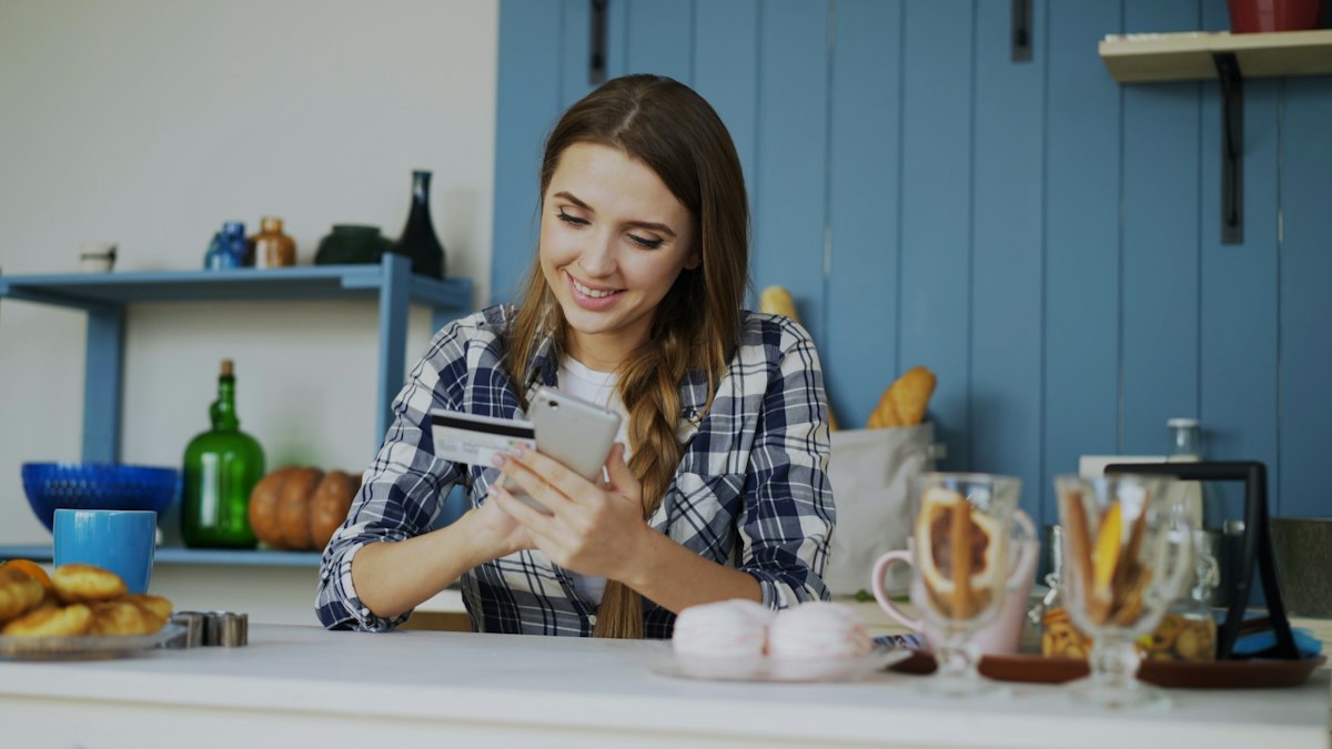 Young woman smiling in her kitchen while paying with a card on her phone